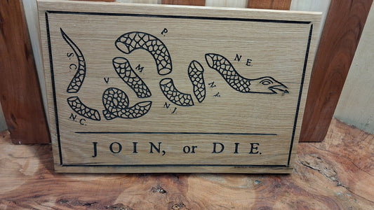 A rectangular wooden sign featuring a black engraved coiled snake illustration with the words 'SC, NC, YS, ME, VA, WV, PA, NY, NJ, NH, VT, VT, CT, RI, MA, ME, VA, WV, PA, NY, NJ, NH, VT, CT, RI, MA, ME' and the phrase 'JOIN, or DIE.' below it, photographed on a wood surface.
